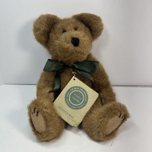 Pode incluir: Um urso de pelúcia marrom com uma fita verde escura amarrada ao redor do pescoço. O urso tem um nariz preto e uma etiqueta que diz "Cranston R. Bean". O urso é um item de colecionador.