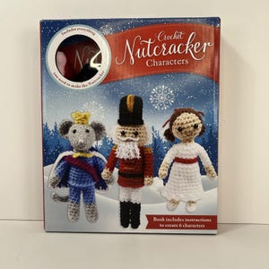 Pode incluir: Uma caixa de kit Crochet Nutcracker Characters com um fundo de floresta de inverno azul. A caixa exibe três bonecos de crochê: um rato em azul, um quebra-nozes em vermelho e uma menina em branco. O texto diz: Crochet Nutcracker Characters.