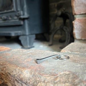 May include: A silver metal horseshoe nail keyring. The keyring features a horseshoe nail design with a small ring for keys. The nail is resting on a textured, reddish-brown brick surface.