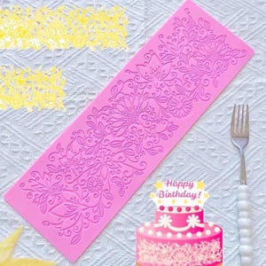 May include: A rectangular pink silicone mould featuring an intricate embossed floral pattern with various flowers, leaves, and swirling vines. The mould is designed for creating decorative fondant or clay impressions and is shown next to a metal fork.