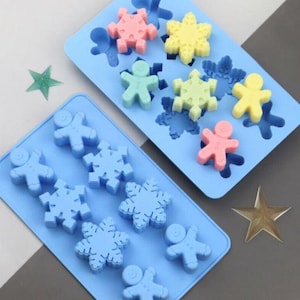 Puede incluir: Dos moldes de silicona azul para hacer caramelos o jabón. Un molde tiene formas de copo de nieve y el otro tiene formas de hombre de jengibre. Los moldes están llenos de caramelos de colores.