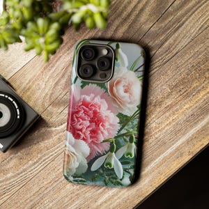 Puede incluir: Una funda para teléfono con un diseño floral con flores rosas, blancas y verdes. La funda tiene un recorte para el objetivo de la cámara y un fondo blanco.