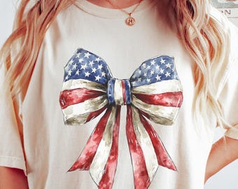 May include: A cream-colored t-shirt with a large watercolor bow printed on the front. The bow is in the colors of the American flag, red, white, and blue.