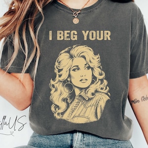May include: A charcoal gray short-sleeve t-shirt featuring a yellow graphic illustration of a woman with voluminous hair and large hoop earrings. The text "I BEG YOUR" is printed in yellow above the illustration. The shirt has a crew neckline.