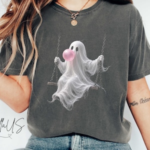Może przedstawiać: Ciemnoszary t-shirt z krótkim rękawem z grafiką przedstawiającą białego ducha siedzącego na huśtawce i puszczającego różową bańkę z gumy do żucia. Koszulka ma okrągły dekolt i luźny krój. W prawym górnym rogu widoczny jest napis Pepper.