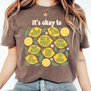 May include: An espresso-coloured t-shirt featuring a graphic print of yellow lemons and green leaves. The text reads: it's okay to BE DIFFERENT, NOT KNOW YET, NOT BE PERFECT, CHANGE YOUR MIND, MAKE MISTAKES, START OVER, ASK FOR HELP, TRY AGAIN.