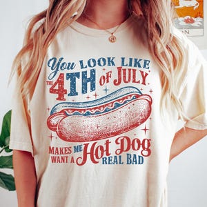 May include: Ivory-colored t-shirt with a patriotic hot dog graphic. The design features the text "You look like the 4th of July" and "Makes me want a Hot Dog real bad" in red and blue. A fun, casual tee for Independence Day.