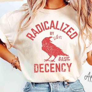 以下が含まれることがあります： アイボリー色のTシャツ。赤色の「RADICALIZED by BASIC DECENCY」の文字と赤いカラスのグラフィックが特徴です。明るい青色のジーンズと組み合わせています。「ApparellaUS」のブランド名が見えます。