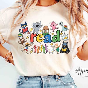May include: An ivory short-sleeve t-shirt featuring a colorful graphic print of various cartoon characters reading books. The central text reads "read" in multi-colored letters. The design includes a cat, elephant, pig, bird, monkey, and crayons.