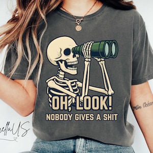 Op de afbeelding: Donkergrijs T-shirt met een skelet dat door een verrekijker kijkt. Het skelet houdt de verrekijker met beide handen vast. De tekst op het shirt luidt "OH LOOK! NOBODY GIVES A SHIT".