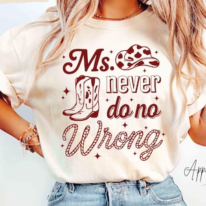 May include: Ivory-colored t-shirt with the text "Ms. Never Do No Wrong" in a maroon font, along with a cowboy hat and boot graphic. The shirt is paired with light blue jeans.