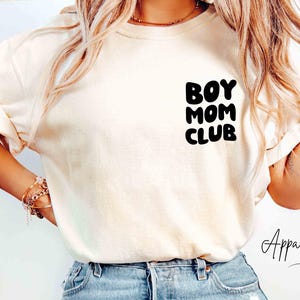 May include: An ivory short-sleeved t-shirt featuring the black text BOY MOM CLUB printed on the upper left chest area. The shirt is paired with light blue denim jeans that have a distressed detail on the upper thigh area of the left leg.