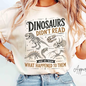May include: An ivory t-shirt featuring a black and brown graphic of various dinosaur skeletons including a T-Rex, Stegosaurus, and Triceratops. The text reads: DINOSAURS DIDN'T READ and we know WHAT HAPPENED TO THEM, along with labels for each fossil.