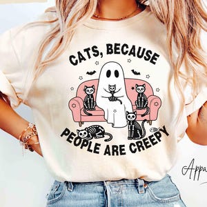 May include: Ivory t-shirt with the slogan "CATS, BECAUSE PEOPLE ARE CREEPY." The design shows a ghost with a cat on a pink sofa, surrounded by black cats and bats. The shirt is worn with light blue jeans.