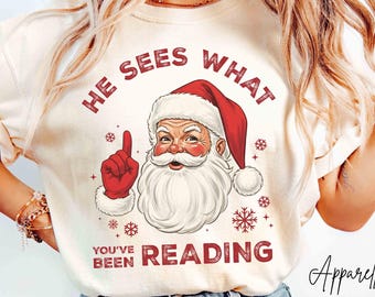 May include: Ivory-colored t-shirt featuring a Santa Claus graphic with the text "HE SEES WHAT YOU'VE BEEN READING" in red. The Santa illustration includes a red hat and gloves. The shirt is paired with light blue jeans.
