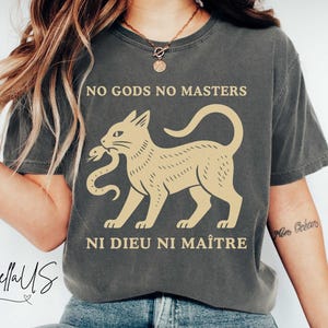Puede incluir: Camiseta gris oscuro con el texto "NO GODS NO MASTERS" sobre un dibujo de un gato beige que sostiene una serpiente. Debajo del dibujo está el texto "NI DIEU NI MAÎTRE". La camiseta es de estilo casual.
