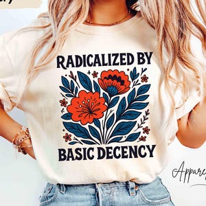 以下が含まれることがあります： アイボリー色のTシャツ。黒字で「RADICALIZED BY BASIC DECENCY」と書かれています。赤い花と青い葉のグラフィック。モデルはライトブルーのジーンズを着用しています。