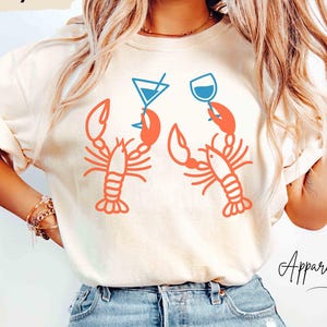 K&ouml;nnte beinhalten: Elfenbeinfarbenes T-Shirt mit einer Grafik von zwei orangefarbenen Hummern, die Cocktailgl&auml;ser halten. Die Gl&auml;ser sind blau. Der Text "ApparellaUS" befindet sich in der unteren rechten Ecke.