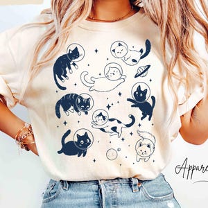 May include: Ivory-colored t-shirt featuring a whimsical design of cats in space helmets, surrounded by stars and planets. The graphic is in navy blue. The text "ApparellaUS" is in the lower right corner.