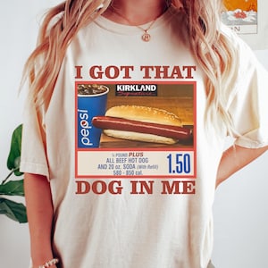 以下が含まれることがあります： 「I GOT THAT DOG IN ME」の文字が入ったアイボリー色のTシャツ。ホットドッグ、ペプシのカップ、カークランドのロゴが描かれています。Tシャツには、ホットドッグが&frac14;ポンドプラスのオールビーフホットドッグと20オンスのソーダであることも記載されています。