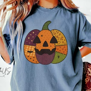 May include: Blue jean colored t-shirt featuring a patchwork pumpkin design with a jack-o'-lantern face. The pumpkin is composed of various patterned fabrics in orange, purple, yellow, and green. The text "ApparellUS" is visible on the left side.