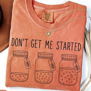 May include: A yam-colored, crew neck t-shirt with the phrase "DON'T GET ME STARTED" printed in black. The shirt features three jar illustrations with leaf tags. A gold necklace and a brown hat are also visible.