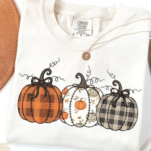 Op de afbeelding: Ivoor kleurig T-shirt met drie decoratieve pompoenen. De pompoenen hebben ruit- en bloempatronen in oranje, wit en grijs. Een gouden ketting met een hanger is over het shirt gedrapeerd.