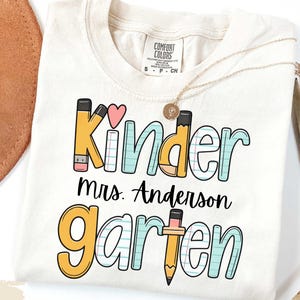 Puede incluir: Camiseta de color marfil con las palabras "Kinder Mrs. Anderson Garten" en una fuente colorida y juguetona. El diseño incluye gráficos de lápices y corazones, perfecto para una maestra de jardín de infancia. Un collar dorado está sobre la camiseta.
