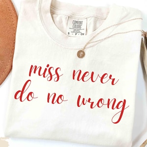 May include: Ivory-colored Comfort Colors t-shirt with the text "miss never do no wrong" in red script. A gold necklace with a pendant rests on the shirt. A brown hat and dried plant are in the background.