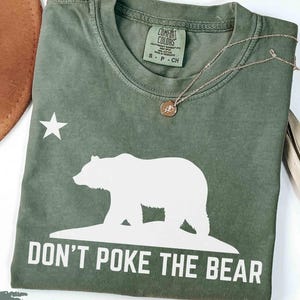 May include: Moss green t-shirt with a white bear graphic, a star, and the text "DON'T POKE THE BEAR." The shirt has a crew neck and short sleeves. A gold necklace with a pendant is draped across the shirt.