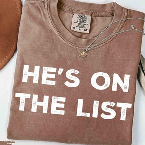 May include: Espresso-coloured t-shirt with the text "HE'S ON THE LIST" in white block letters. The shirt has a crew neck and short sleeves. A silver necklace with a small pendant is draped across the shirt. The shirt is folded on a white surface.