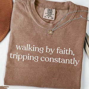 May include: Espresso-colored t-shirt with the text "walking by faith, tripping constantly" in white. The shirt has a crew neck and a Comfort Colors tag. A silver necklace with a pendant is draped on the shirt.