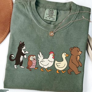 以下が含まれることがあります： モスグリーンのTシャツ。猫、フクロウ、鶏、アヒル、クマの漫画の動物のデザイン。クルーネックとComfort Colorsのタグが付いています。ペンダント付きのゴールドネックレスがTシャツにかけられています。
