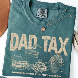 May include: Blue spruce t-shirt with the words "DAD TAX" in a distressed font, along with illustrations of a burger, fries, soda, and ice cream cone. The text "MAKING SURE IT'S NOT POISON" is below the graphic. A gold necklace is draped on the shirt.