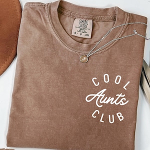 May include: A brown t-shirt with the text "COOL AUNTS CLUB" in white lettering. The shirt is made of a soft, comfortable material and has a relaxed fit.