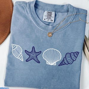 Könnte beinhalten: Ein T-Shirt in Jeansblau mit einem weißen Muschel-, Seestern- und Muschelmotiv. Der Text "Comfort Colors" ist auf dem Etikett sichtbar.