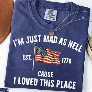 Può includere: T-shirt blu navy con la scritta "I'M JUST MAD AS HELL CAUSE I LOVED THIS PLACE" e una bandiera americana. La maglietta include l'anno "1776" e il marchio "Comfort Colors". Una collana d'oro è drappeggiata sulla maglietta.