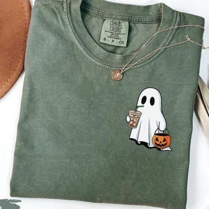 Könnte beinhalten: Ein moosgrünes T-Shirt mit einem Cartoon-Geist, der einen Kaffee und eine Kürbislaterne hält. Das Shirt hat einen Rundhalsausschnitt und ein kleines Etikett am Kragen. Eine goldene Halskette ist über dem Shirt drapiert.