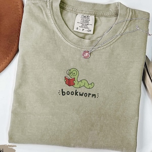 May include: A khaki-coloured short-sleeve t-shirt featuring an embroidered graphic of a green worm wearing glasses and reading a red book. Below the graphic, the word bookworm is embroidered in black. The shirt has a Comfort Colors brand label.