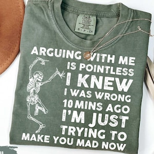 May include: Moss green t-shirt with the text "ARGUING WITH ME IS POINTLESS I KNEW I WAS WRONG 10 MINS AGO I'M JUST TRYING TO MAKE YOU MAD NOW" and a skeleton graphic. A gold necklace is draped across the shirt.