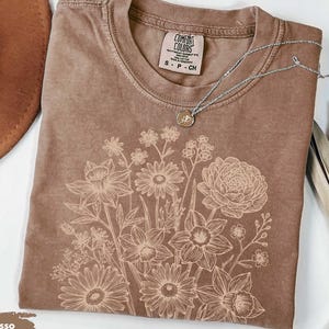 Pode incluir: Camiseta cor de caf&eacute; com um design floral. A camiseta tem gola redonda e mangas curtas. Um colar de prata com um pingente est&aacute; drapeado sobre a camiseta. A camiseta est&aacute; rotulada como "Comfort Colors".