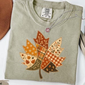 Puede incluir: Una camiseta de manga corta color caqui con un gráfico central grande de una hoja de arce compuesta por varios patrones de tela estilo patchwork en tonos naranja, marrón, crema y verde, con detalles de costura visibles en los bordes.