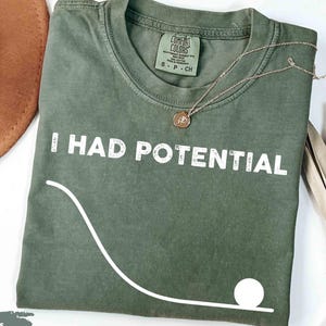 Könnte beinhalten: Ein moosgrünes T-Shirt mit Rundhalsausschnitt und dem weißen Schriftzug I HAD POTENTIAL auf der Brust. Unter dem Text befindet sich eine weiße grafische Darstellung einer abfallenden Kurve, die in einem Kreis als Ball endet.