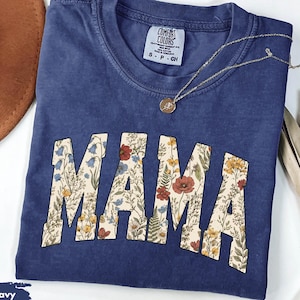 May include: A navy blue short-sleeve t-shirt with a crew neckline. The word MAMA is printed across the chest in large block letters filled with a colorful floral pattern featuring red, yellow, and blue wildflowers on a cream-colored background.