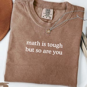 May include: An espresso-colored short-sleeve t-shirt with a crew neckline. The shirt features the white printed text "math is tough but so are you" centered on the chest. A Comfort Colors brand label is visible inside the collar area.