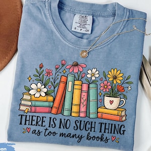 May include: A blue jean coloured t-shirt featuring a graphic of colourful stacked books and flowers. The text reads, There is no such thing as too many books. The shirt has a crew neckline and a Comfort Colors brand label on the inside collar.