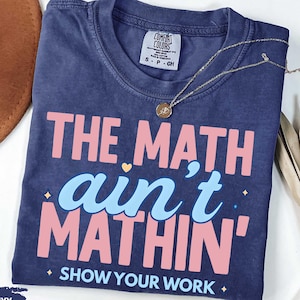 May include: True navy blue t-shirt with the text "THE MATH ain't MATHIN' SHOW YOUR WORK" in pink and light blue. A gold necklace with a pendant is on the shirt. A brown hat and dried plant are in the background.