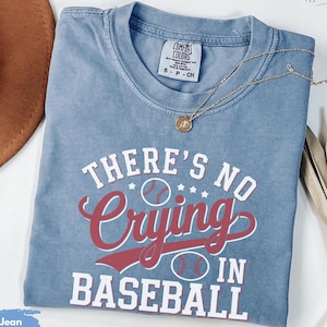 May include: A blue jean t-shirt with the text "THERE'S NO Crying IN BASEBALL" in white and red lettering. The shirt features a baseball graphic.