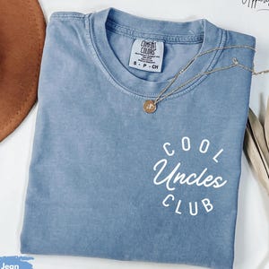 May include: A blue jean t-shirt with the text "Cool Uncles Club" in white lettering. The shirt is folded and laid flat on a surface.