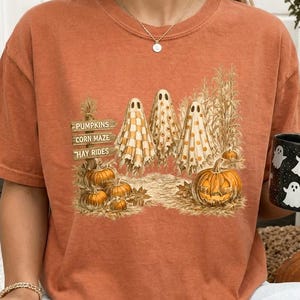 Pode incluir: Uma t-shirt cor de laranja queimado com um gráfico de três fantasmas cobertos por lençóis estampados num campo com abóboras e pés de milho. O gráfico inclui placas de madeira com as palavras PUMPKINS, CORN MAZE e HAY RIDES, além de abóboras.